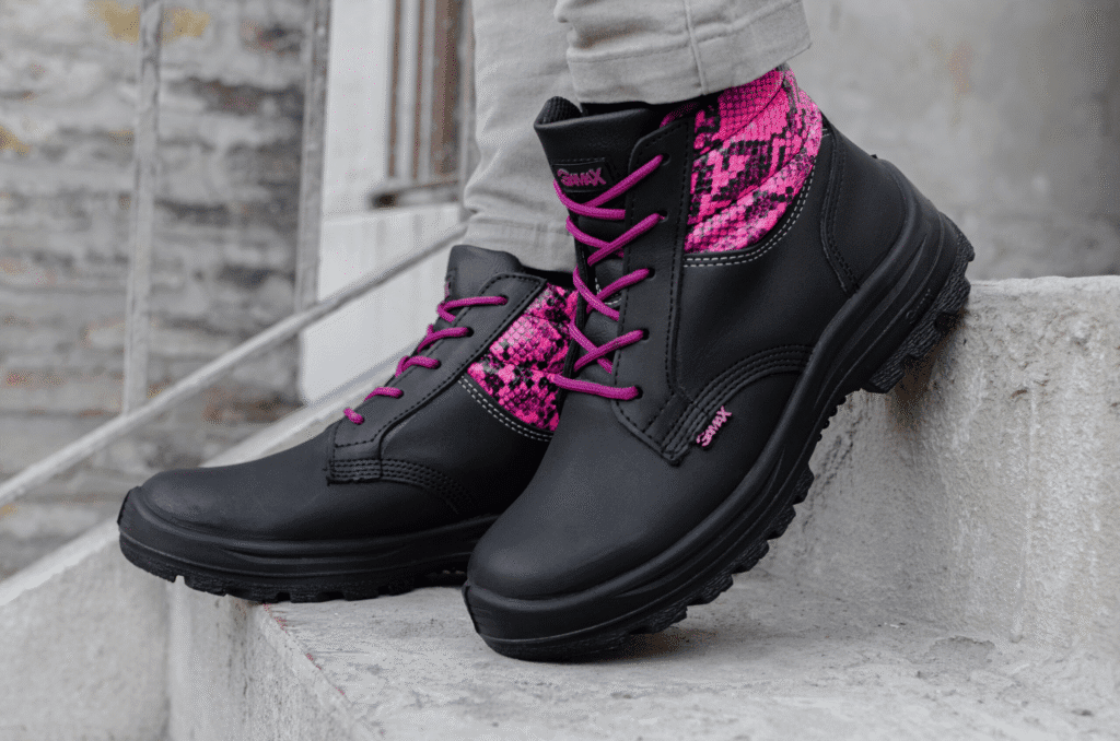 Mujer usando un par de botines de seguridad GamaX, modelo 103FA-113FP, de color fucsia y negro con un estampado de pitón en la caña y cordones fucsia.